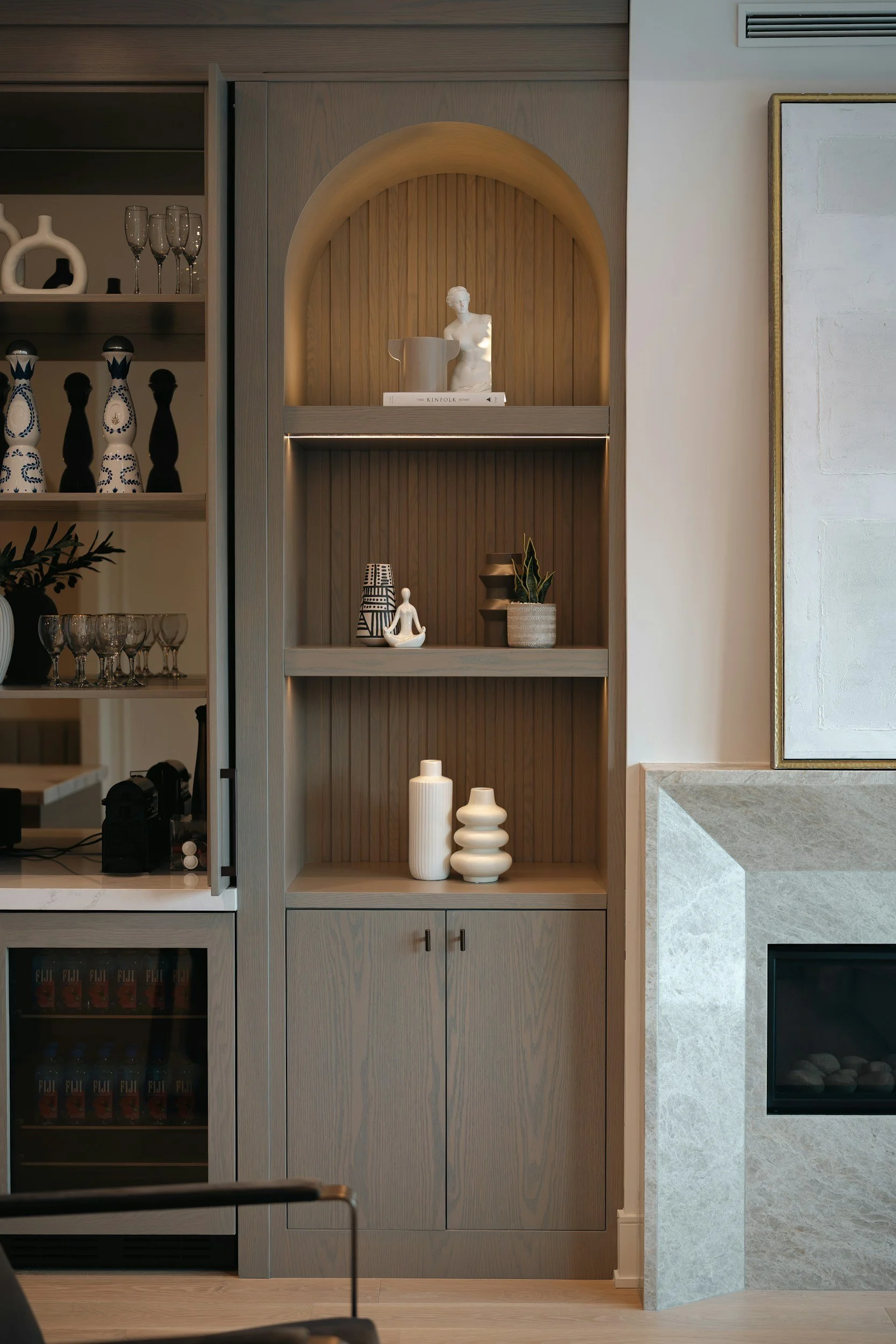 Modern kitchen display with open shelving. Top shelf features a white bust sculpture and a white planter. Middle shelf has a potted plant, vases, and a small sculpture. Bottom shelf contains vases and ceramic decorative objects. To the right, there's a marble counter and a section of a light-colored textured wall.