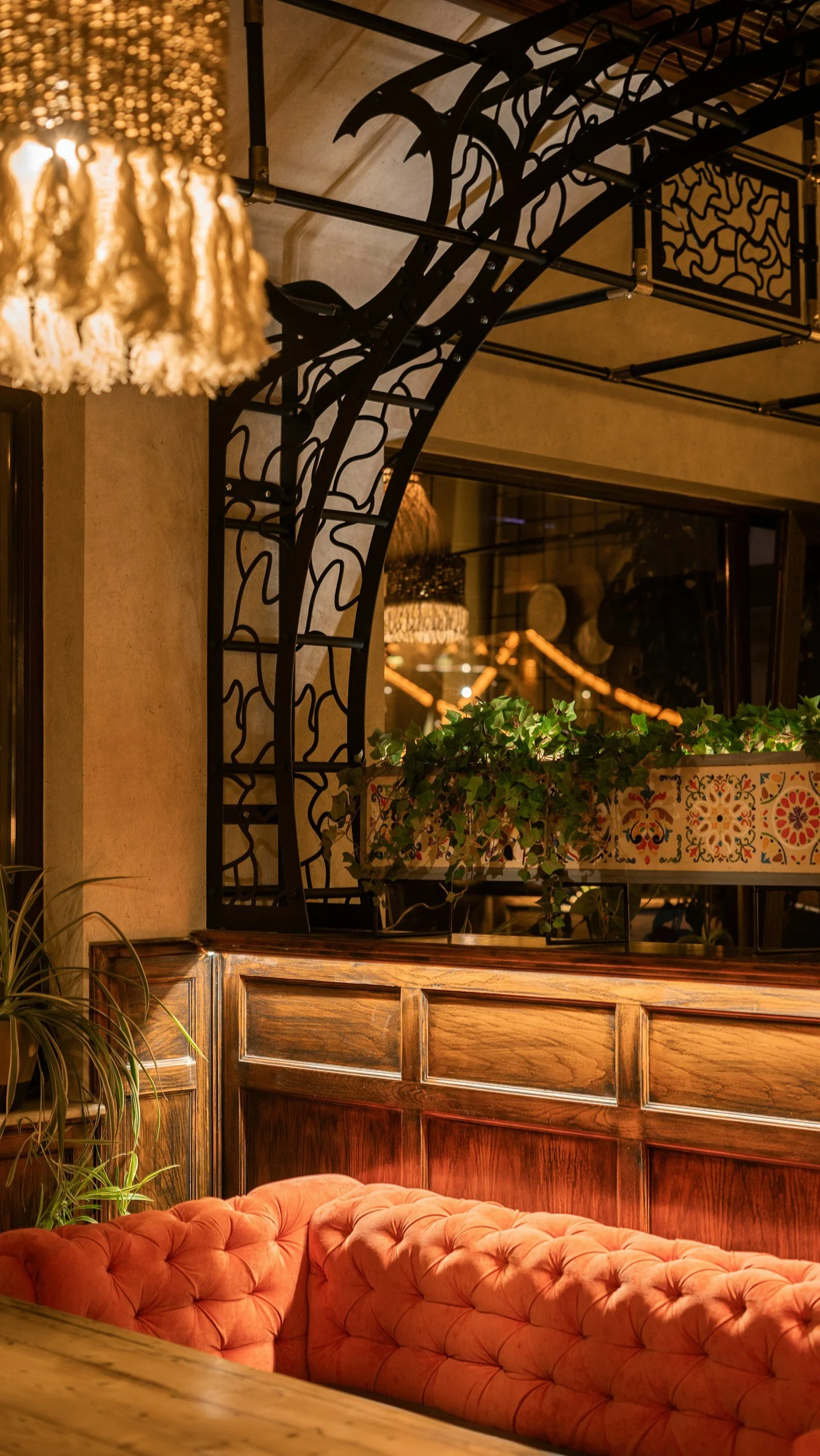 Warmly lit interior with a plush, tufted coral sofa in the foreground, wooden paneling on the wall, decorative iron balcony railing above, and greenery in a colorful planter. A blurry chandelier is visible in the background.