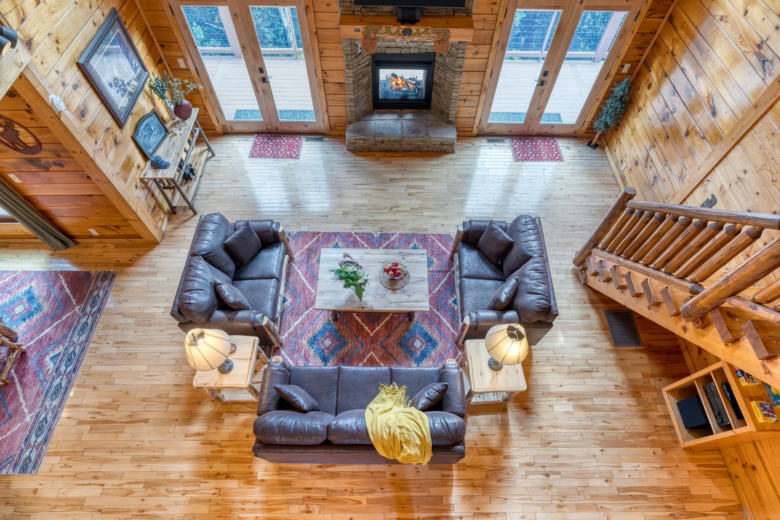 View of a cozy living room with a wood-paneled interior, a central fireplace with a fire burning, plush leather sofas arranged around a wooden coffee table with a bowl of apples and flowers, and large glass doors leading to a deck outside.