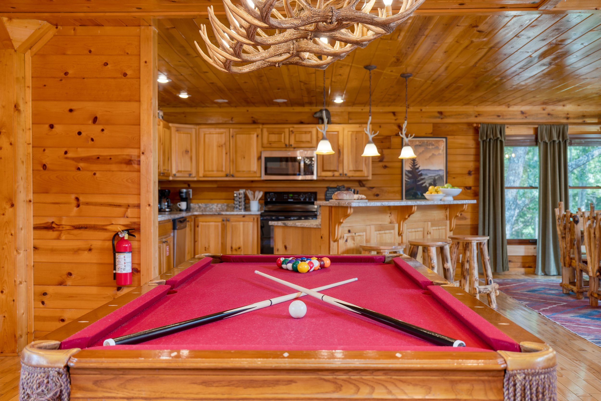 View of a wooden cabin interior with a red pool table in the foreground and a kitchen in the background, featuring wooden cabinets, hanging light fixtures with antler design, and a window with curtains.