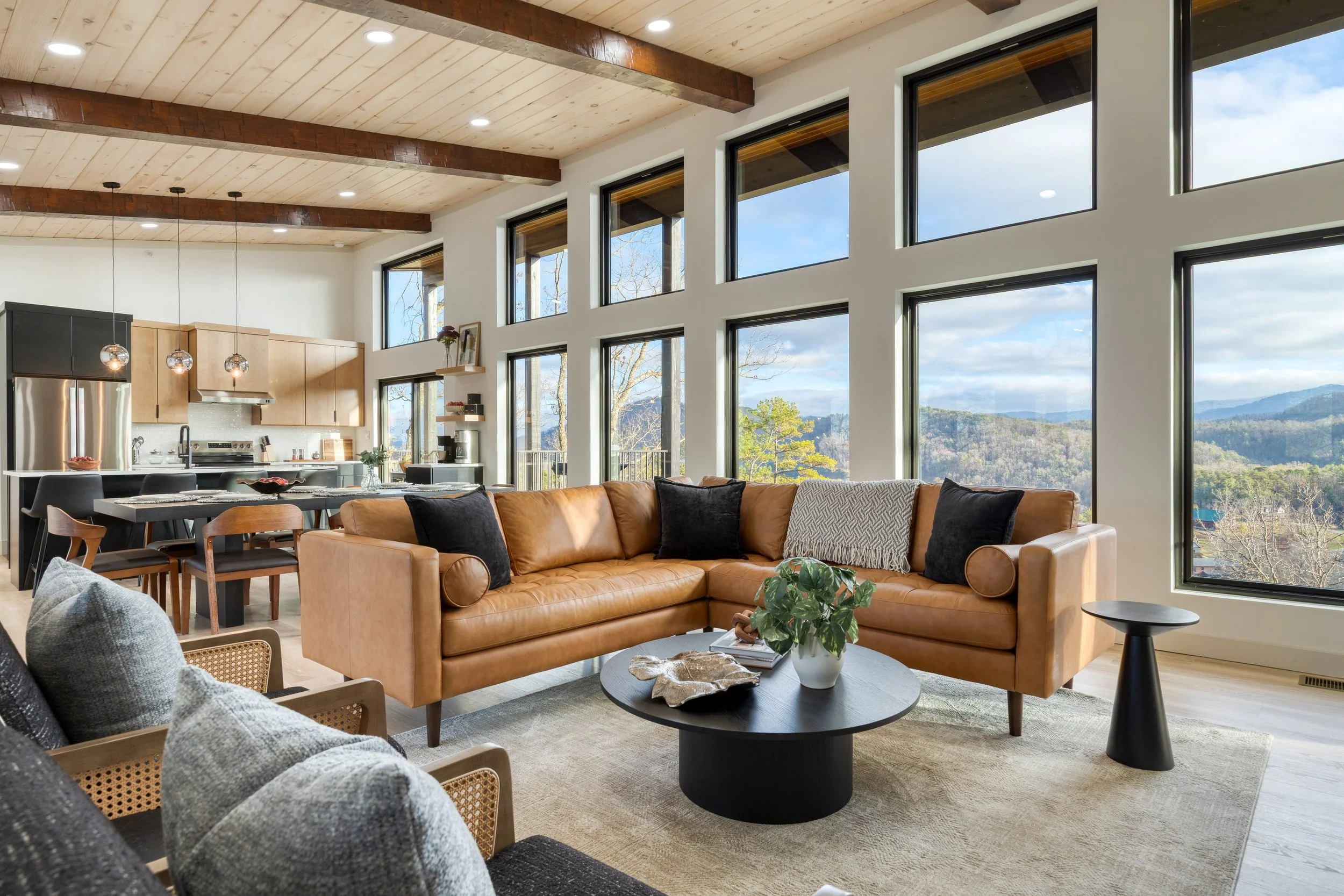 Open living room with large windows overlooking mountains, tan couches, black round coffee table, and wooden ceiling with beams.