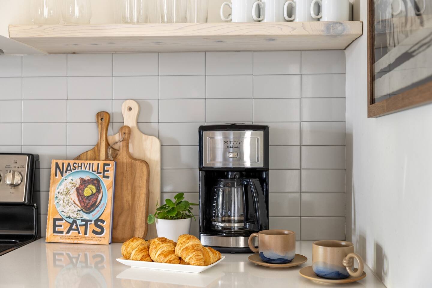 Kitchen countertop with a coffee maker, two ceramic cups on saucers, a plate of croissants, a potted plant, a wooden cutting board, and a Nashville EATS magazine.