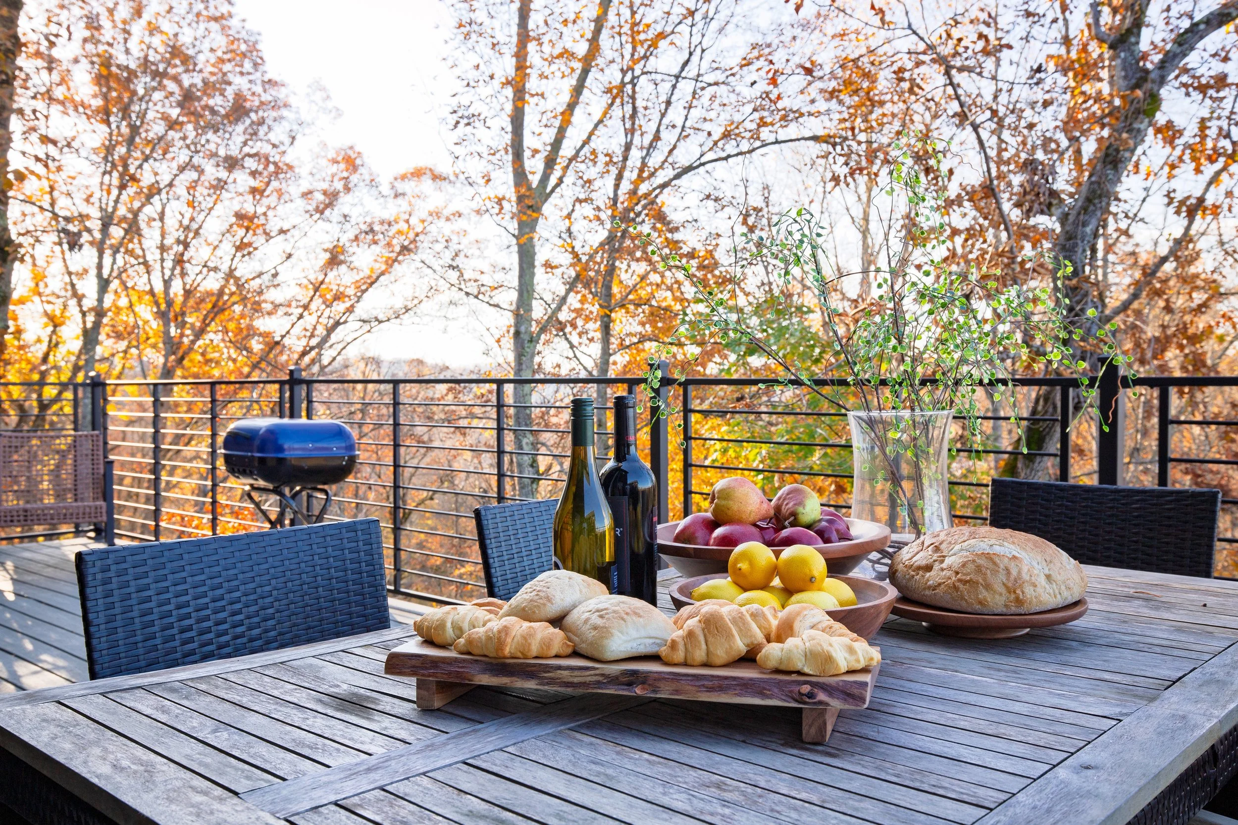 An outdoor wooden deck with a table set for a meal, featuring a bowl of apples, a loaf of bread, and croissants. There are two wine bottles, a glass jar with a green plant, and a small black grill. The background shows a black metal railing and trees