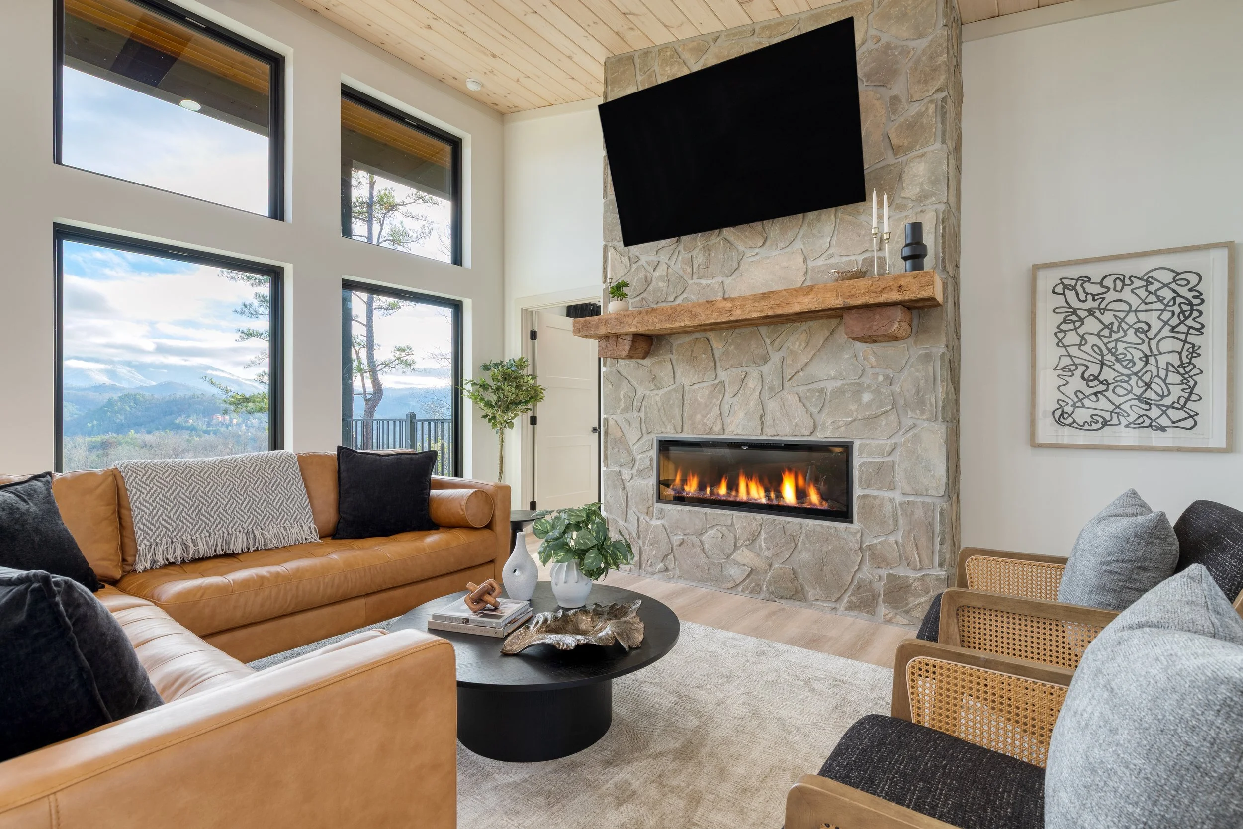 Living room with large windows, a stone fireplace with a mounted TV, a tan leather sofa with black and white cushions, a black coffee table with books and decor, and abstract artwork on the wall.