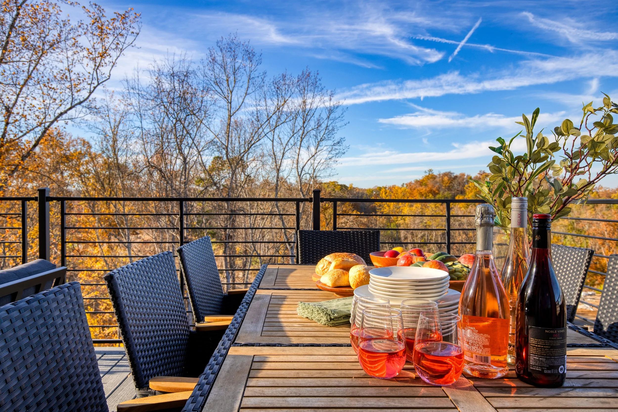 An outdoor dining table on a balcony with a view of trees with autumn leaves and a blue sky with wispy clouds. The table has wine bottles, glasses filled with rosé wine, bread rolls, a bowl of apples, and a vase with green foliage. There are black ch