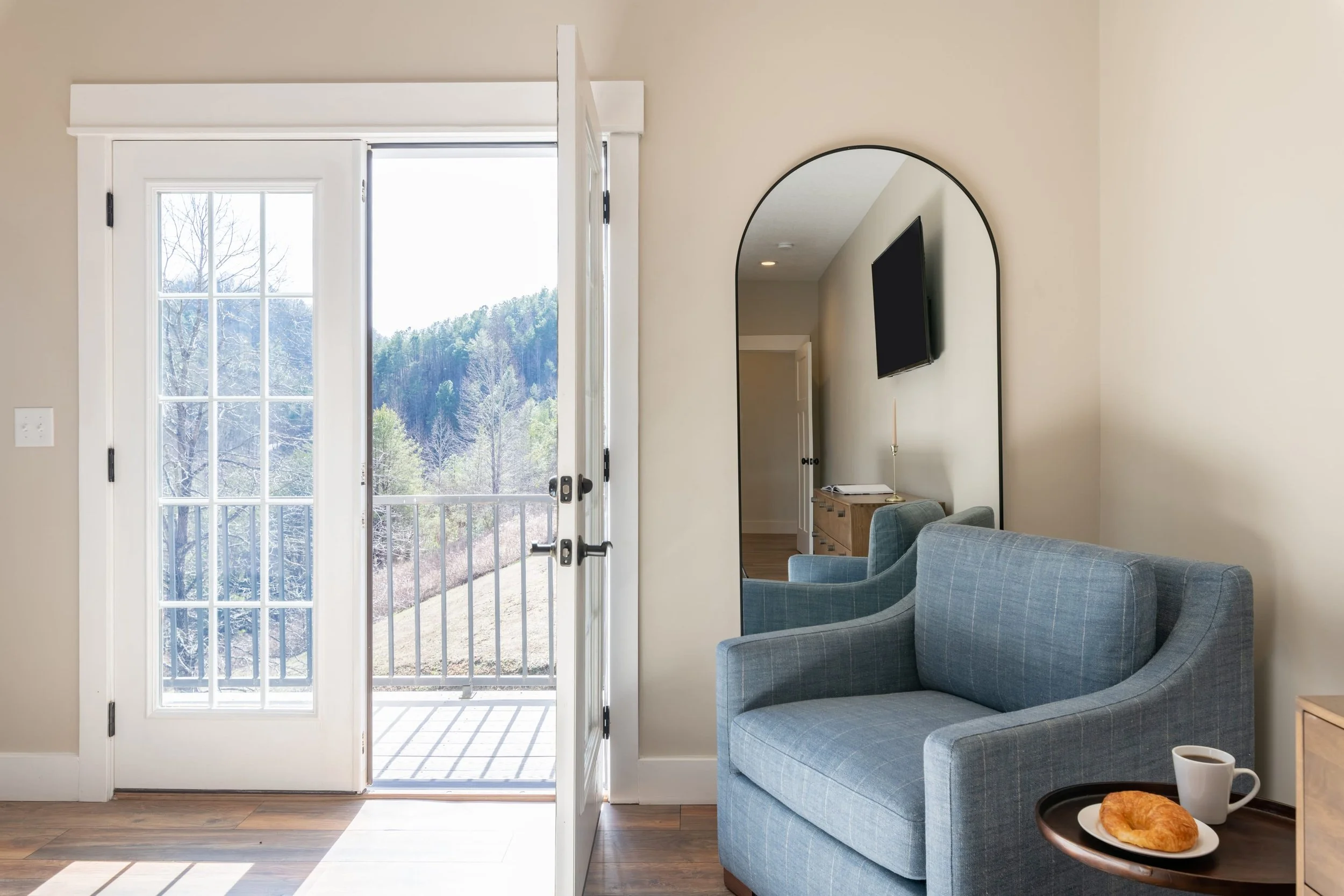 View of a room with a glass door leading to a balcony and a scenic outdoor landscape of trees and hills. Inside, there is a blue checkered armchair with a small table holding a breakfast plate with a croissant and a coffee mug. A flat-screen TV is mo