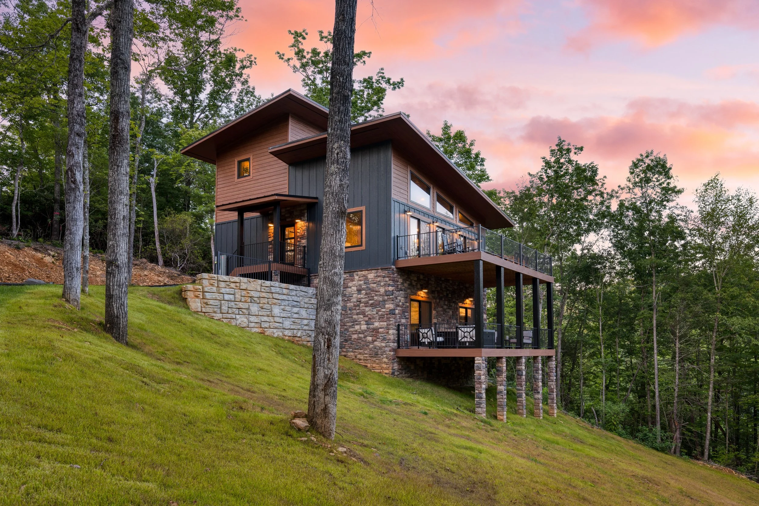 Modern multi-story house with large windows, surrounded by trees on a grassy hillside at sunset