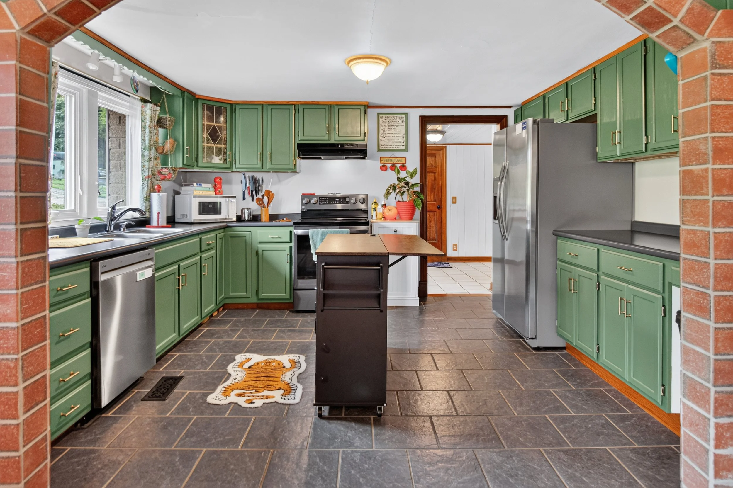 view of kitchen through archway. green cabinets, grey tile floor, orange tiger rug, black rolling island, stainless steel dishwasher and refrigerator, white ceiling, doorway into back room.