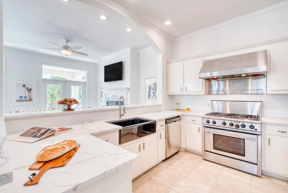 Modern white kitchen with stainless steel stove, hood, dishwasher, and black sink, with marble countertops and bright lighting.