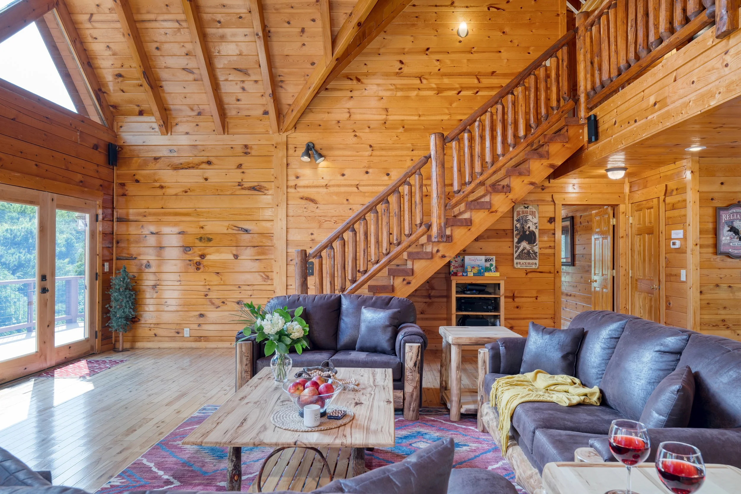 Interior of a wooden cabin living room with high vaulted ceiling, large windows, a cozy gray sectional sofa, a coffee table with a bowl of apples, and wall decorations.