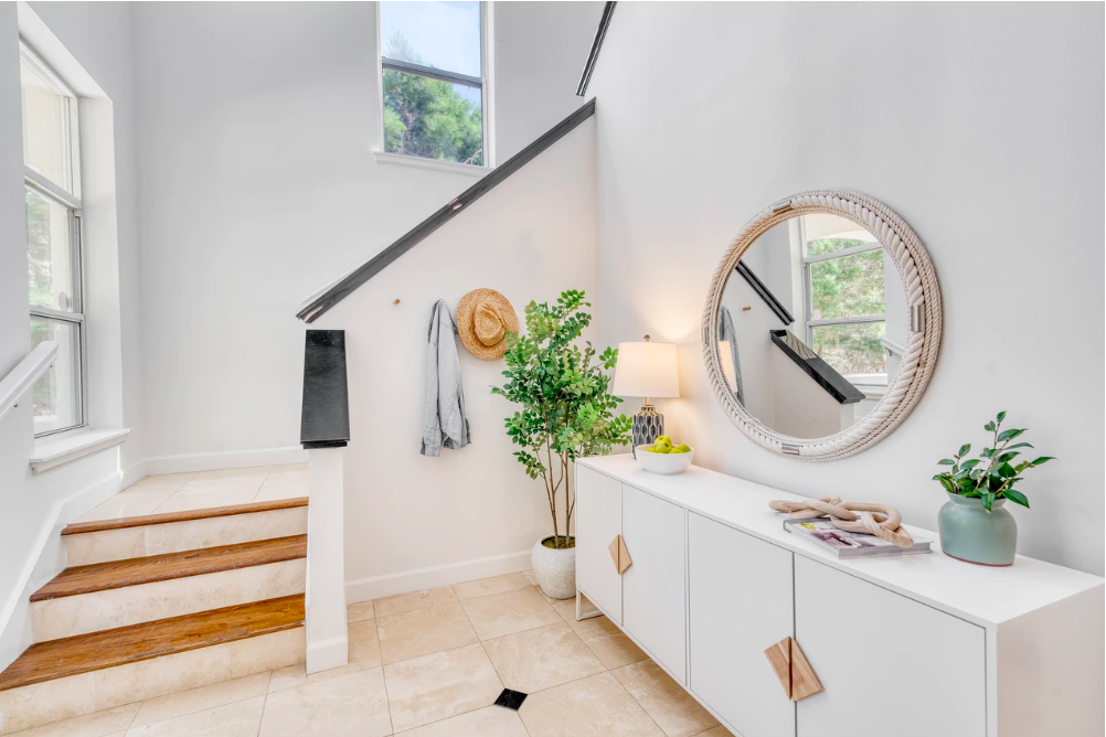 A bright entryway or hallway with stairs on the left, a white sideboard on the right, a large round mirror, a lamp, a potted plant, and decorative items, with a window showing greenery outside.