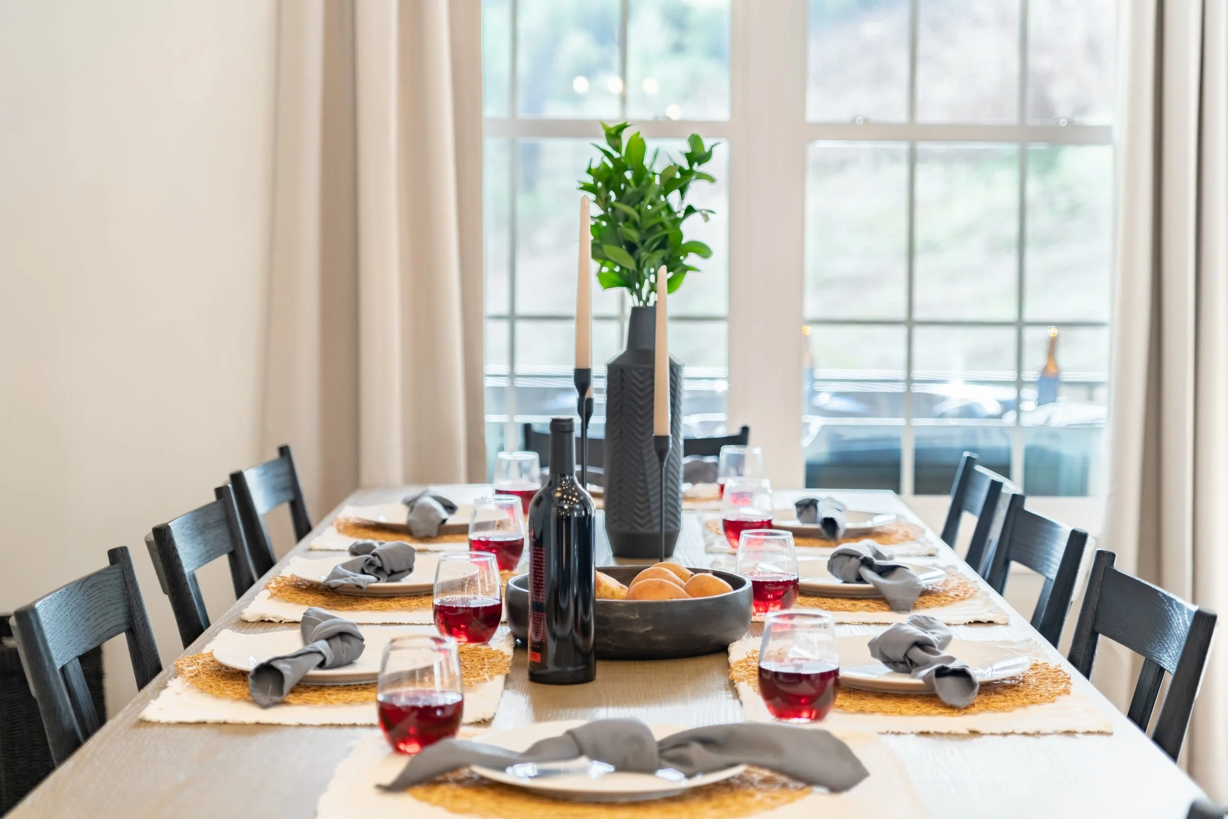 A dining table set for a meal with wine glasses, napkins, and a centerpiece with green plant and candles, in front of large windows with white curtains.