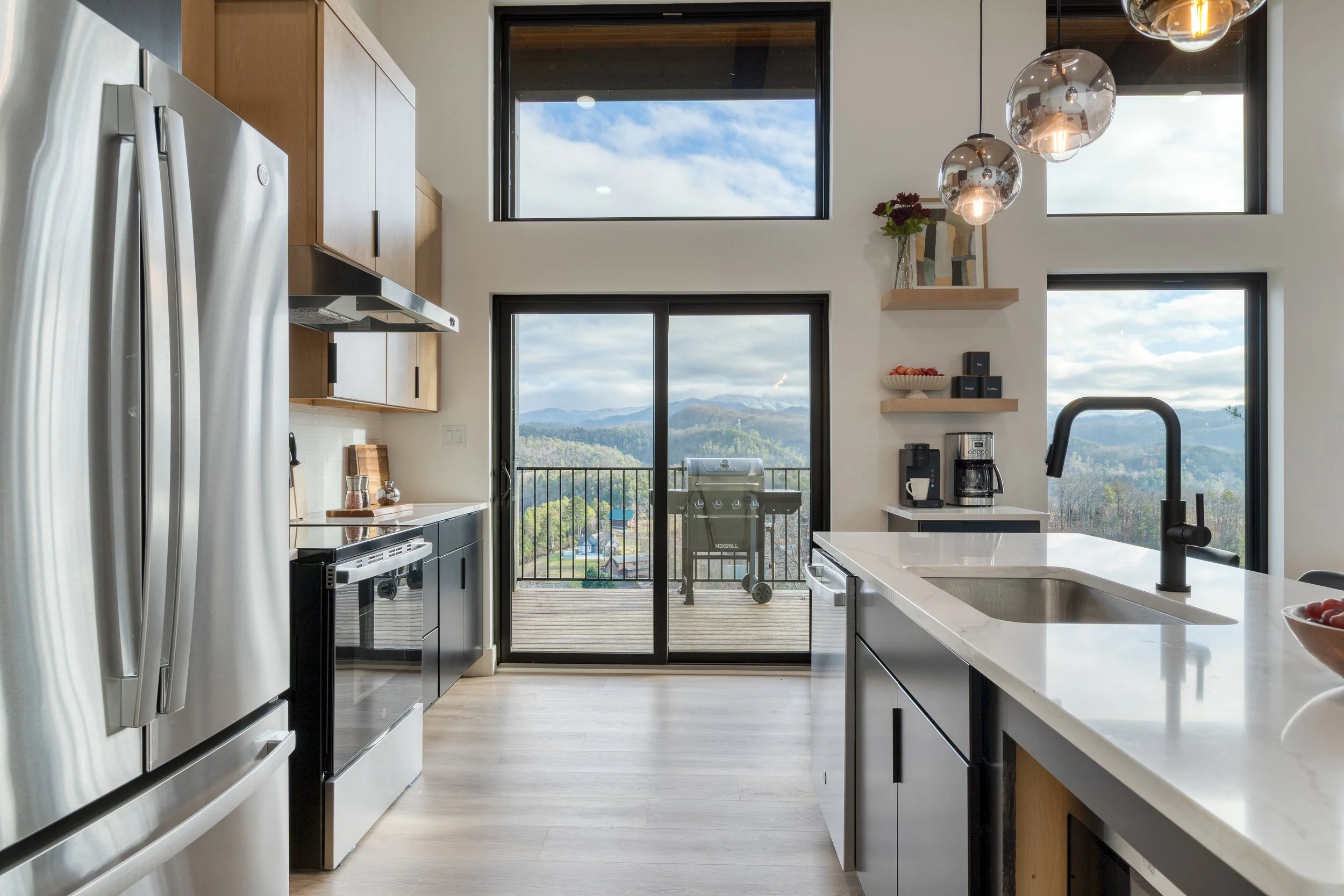 Modern kitchen with large windows showing a mountain view, stainless steel appliances, wooden cabinets, and a white countertop with a black faucet.