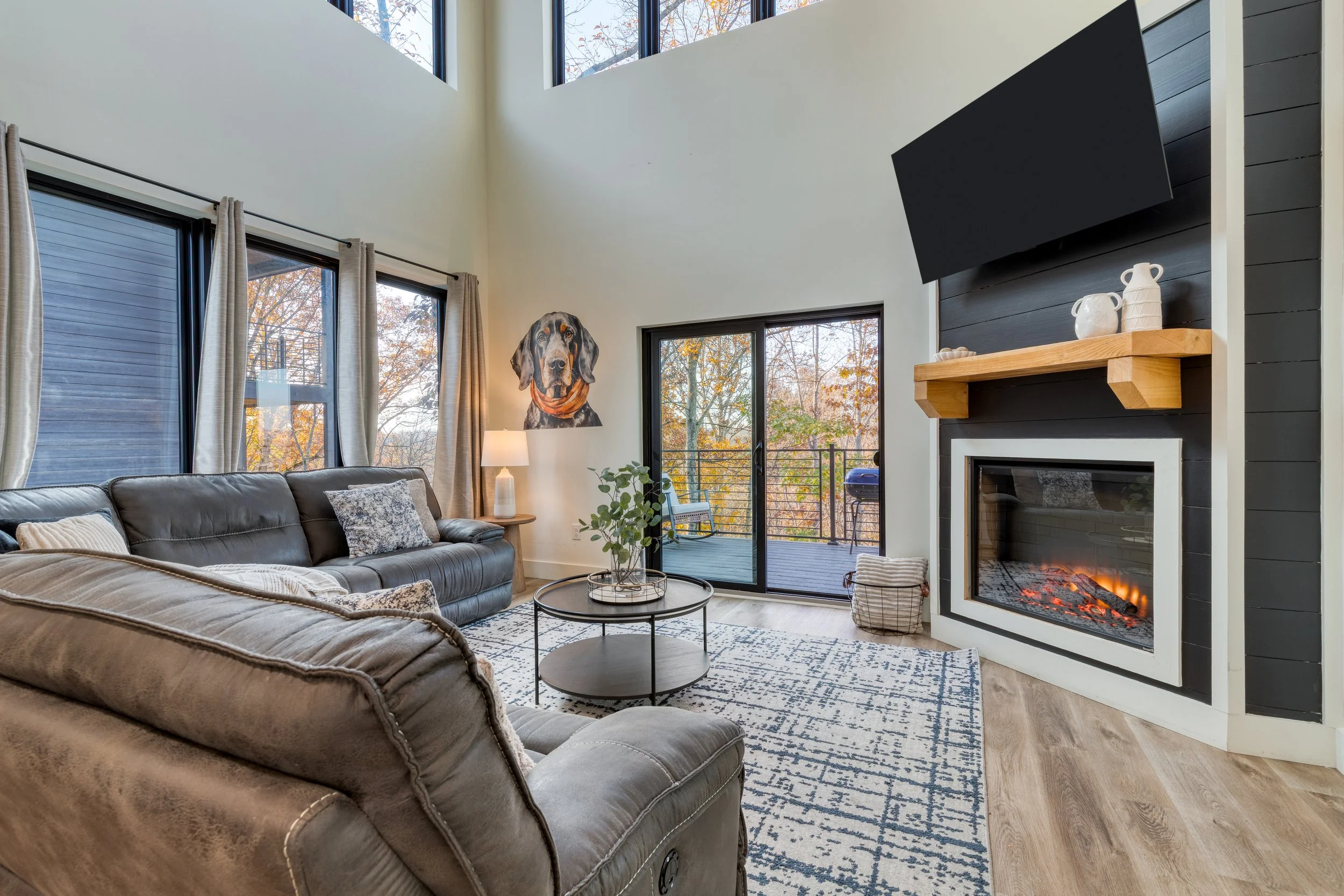 Living room with large windows revealing trees with fall foliage, a painting of a dog on the wall, a gray sofa with pillows, a round coffee table with plants, a patterned rug, a fireplace with a wooden mantle decorated with vases, and a wall-mounted 