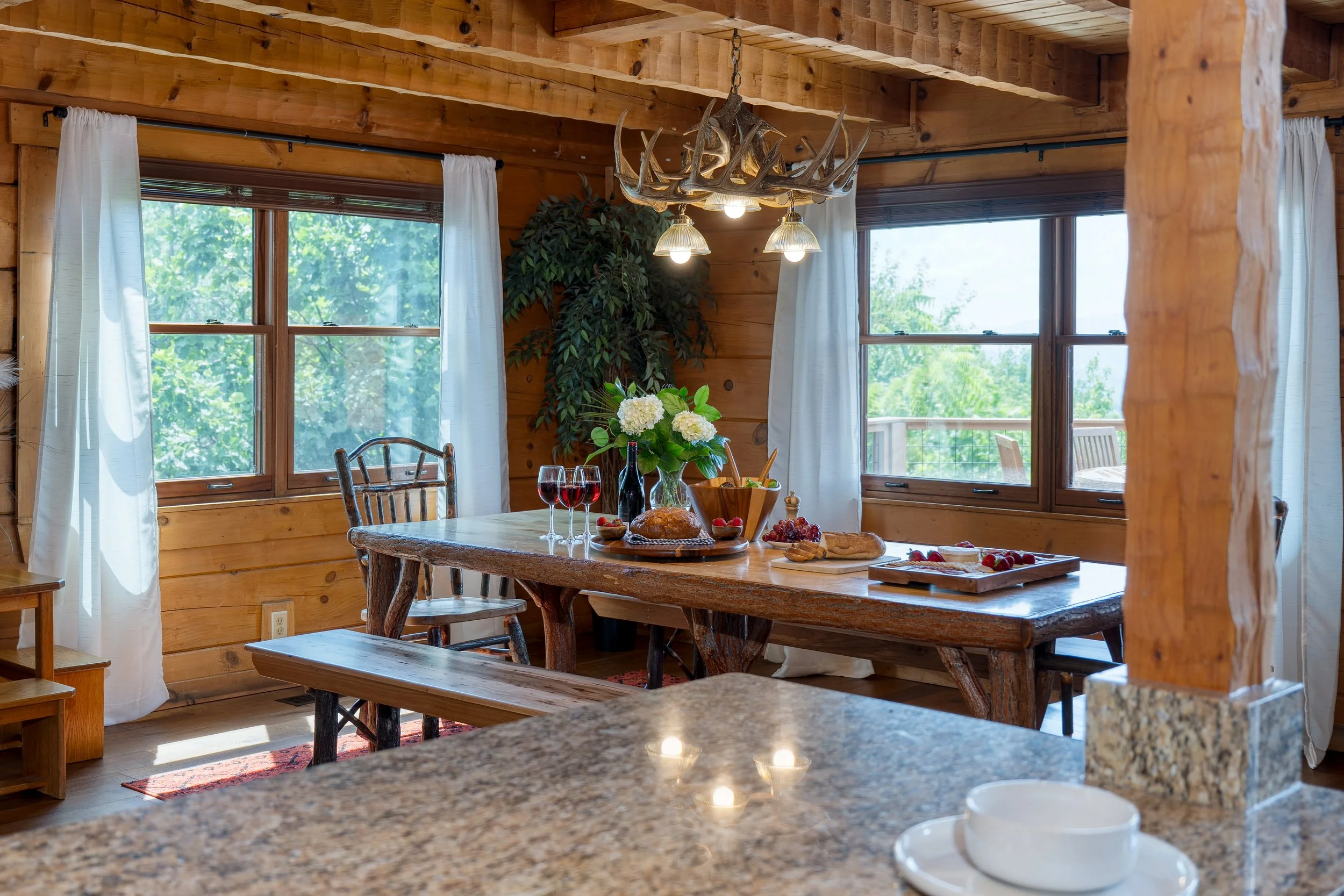 A rustic dining room with large windows, a wooden table set for breakfast with wine, bread, and fruit, and a chandelier made of antlers hanging above.