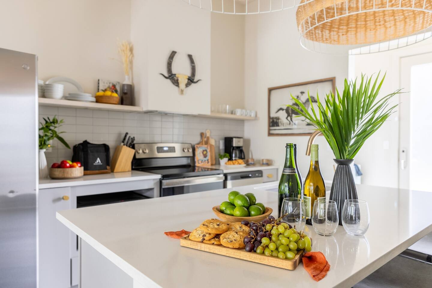 Modern kitchen with a white countertop, green grapes, limes, wine bottles, glasses, a potted plant, and cookies on a tray.