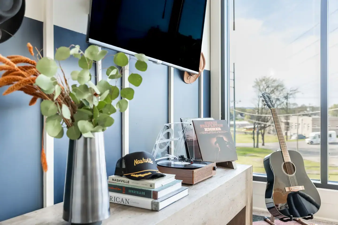 A modern interior with a blue wall, a flat-screen TV, a city view through large windows, a guitar, a vase with eucalyptus leaves and orange flowers, vinyl records, hats, and books on Nashville and Australian topics.
