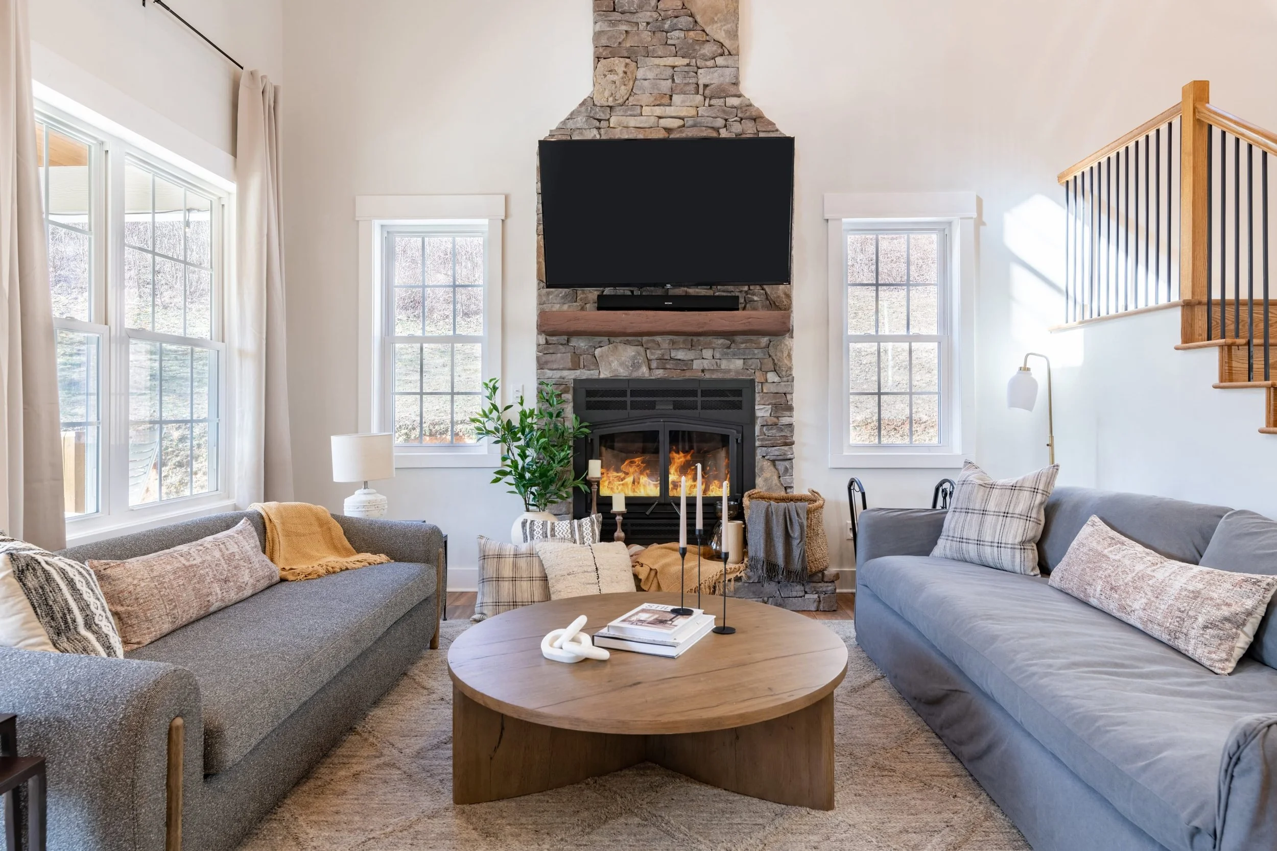 Living room with two sofas, a stone fireplace with a mounted TV, large windows, a wooden coffee table, and various decorative pillows and objects.