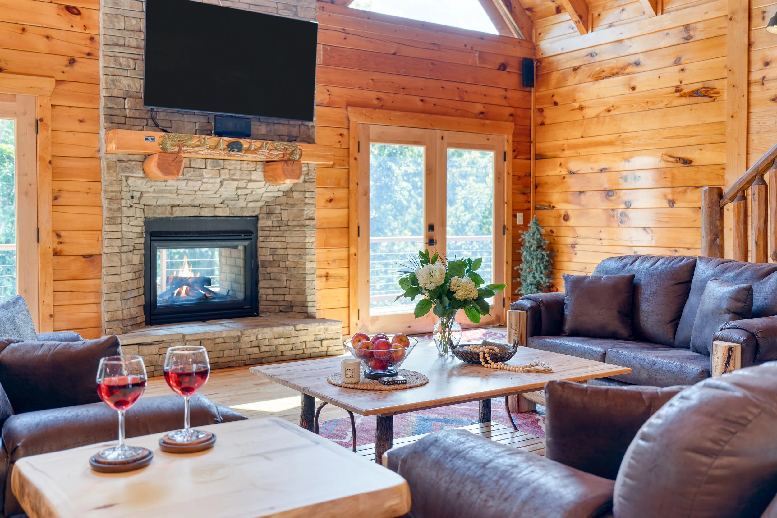 Living room with wood-paneled walls, a stone fireplace with a television above, large windows, a sofa, a coffee table with apples, a vase with flowers, and glasses of red wine.