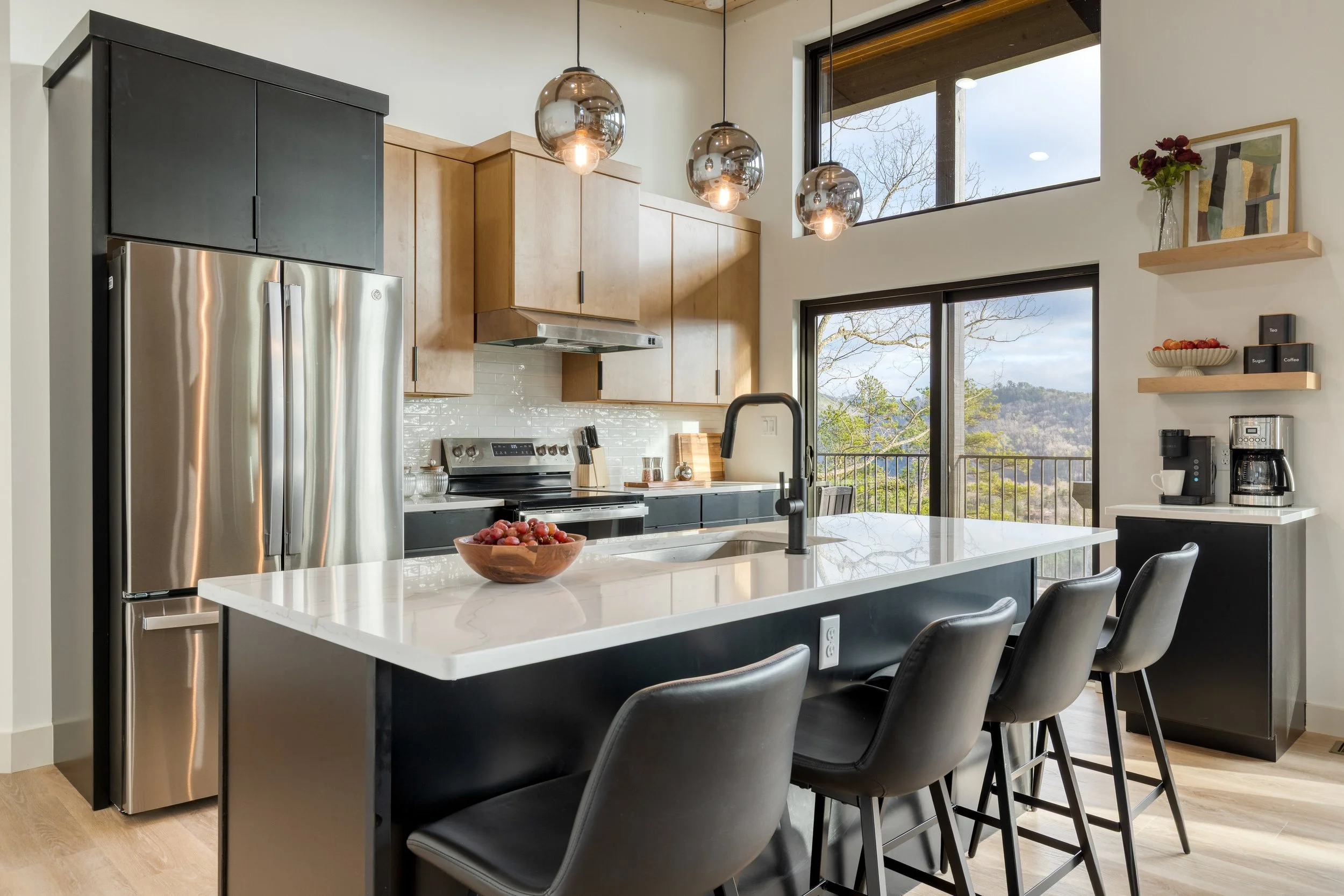 Modern kitchen with a white island counter, black cabinets, stainless steel refrigerator, wooden upper cabinets, pendant lights, large windows, and a view of trees and hills outside.