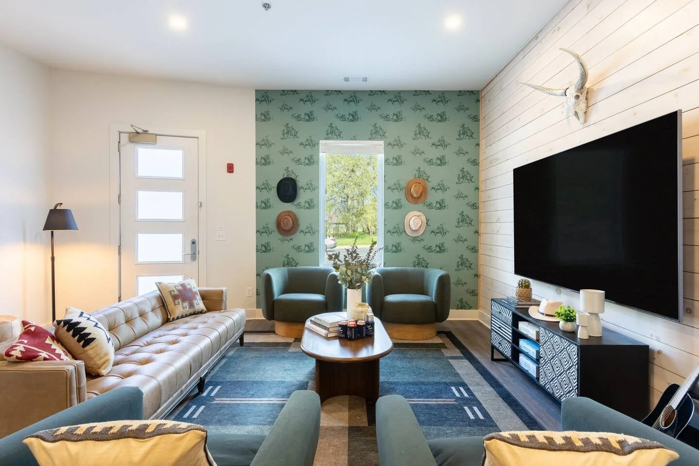 Living room with a beige leather couch, two green armchairs, a wooden coffee table, and a large flat-screen TV on a white wooden wall. Decor includes a mounted animal skull, a patterned black and white TV stand, and sunlight coming through a window w