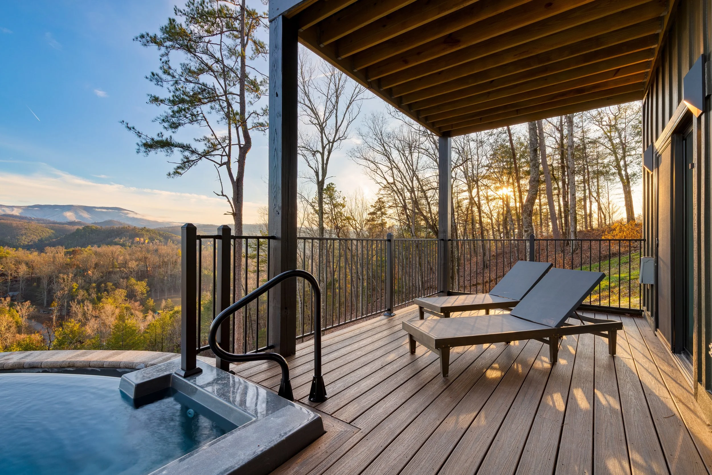 A wooden deck with two lounge chairs overlooking a scenic mountain view at sunset, with trees and a distant mountain range.