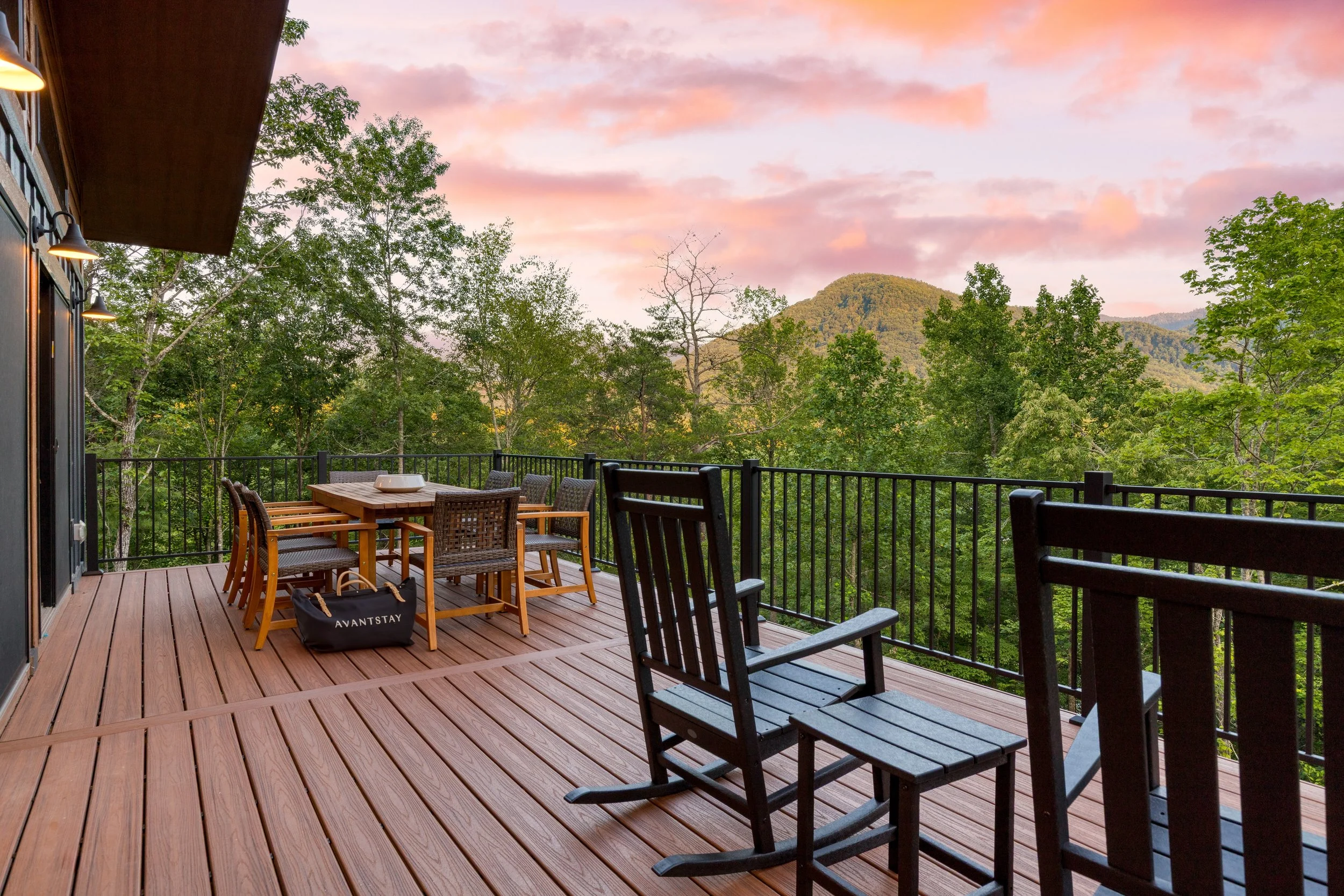 A wooden outdoor balcony with black railing, outdoor dining table with chairs, surrounded by green trees, mountain in the background, pink and purple sky at sunset.