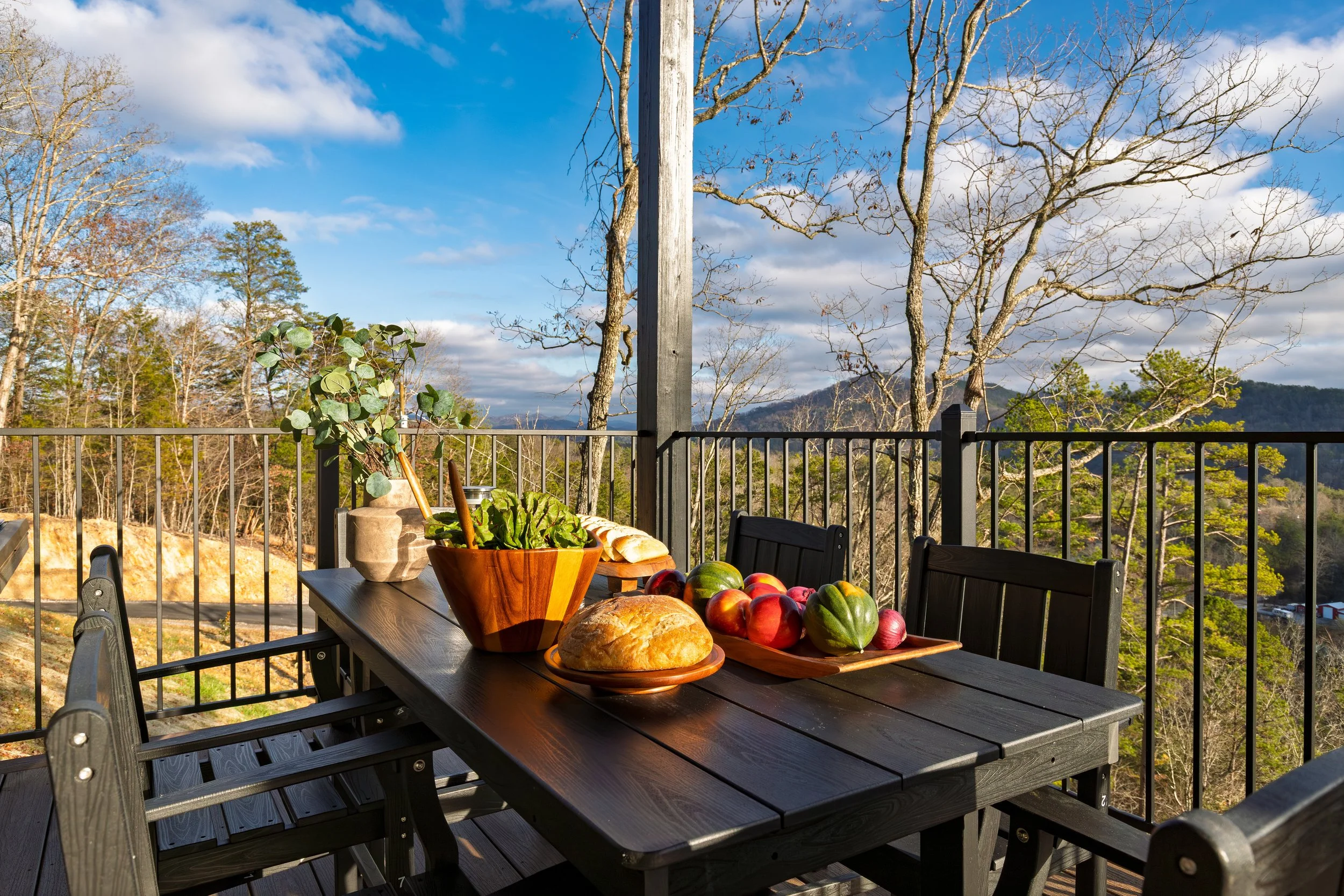 Outdoor patio table set with bread, apples, leafy greens, and decorative plant, overlooking a scenic view of trees and mountains on a bright sunny day.