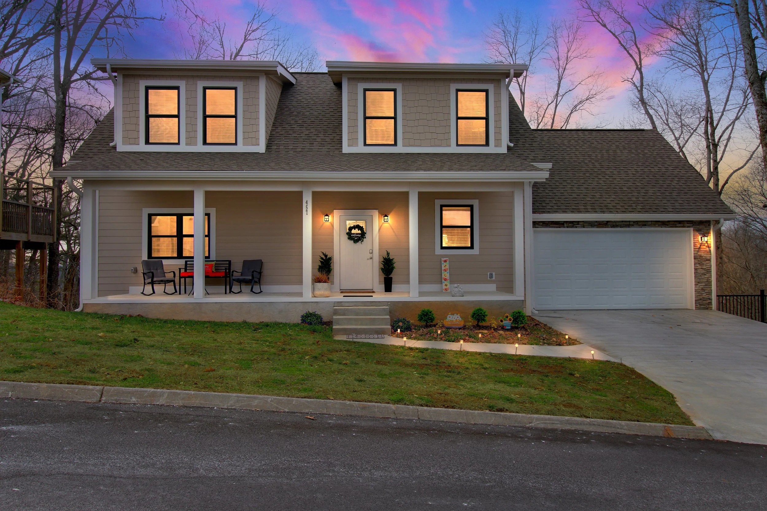 A two-story house with tan siding and a gabled roof, lit from the inside, with a front porch area decorated with potted plants, outdoor chairs, and a small festive wreath on the door. The house is surrounded by leafless trees and a grassy yard, with 