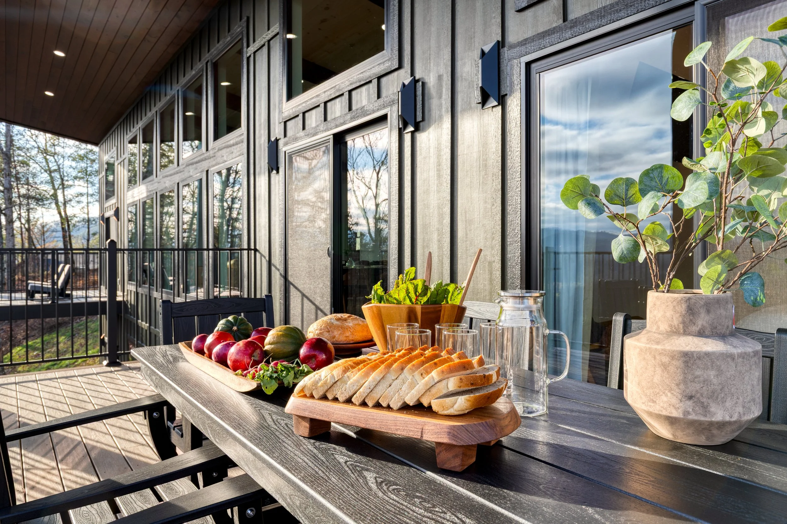Outdoor patio with a table set for a meal, featuring a tray of sliced bread, apples, a pumpkin, and a plant in a pot, with a modern house and trees in the background.