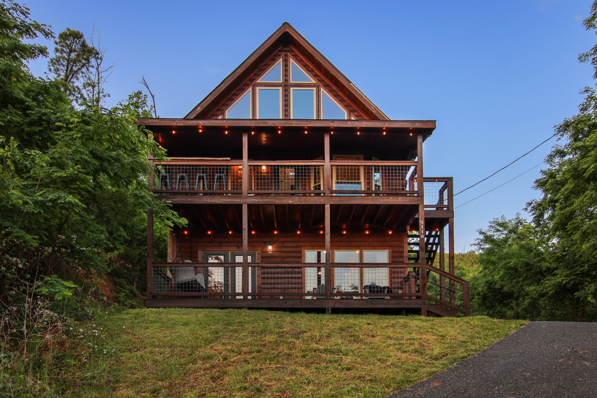 A multi-story wooden house with large windows and a balcony, surrounded by greenery, under a clear blue sky, with string lights on the balcony.