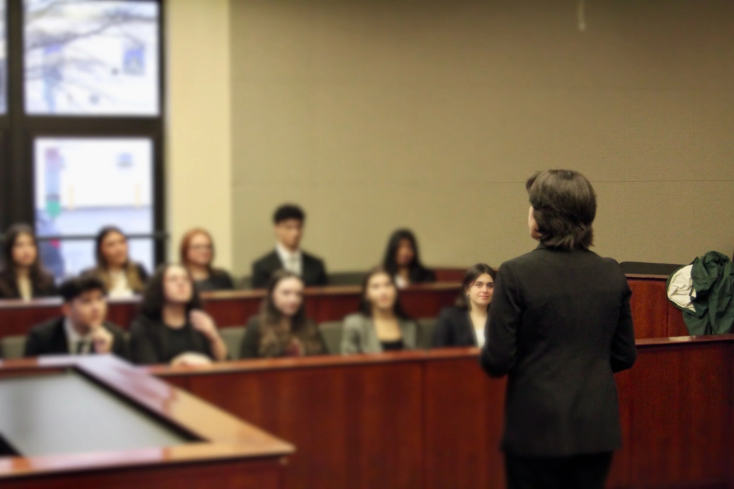 A teacher or speaker with shoulder-length brown hair, facing a group of students in a classroom or lecture hall with beige walls and large windows, some students are paying close attention, others are slightly distracted.
