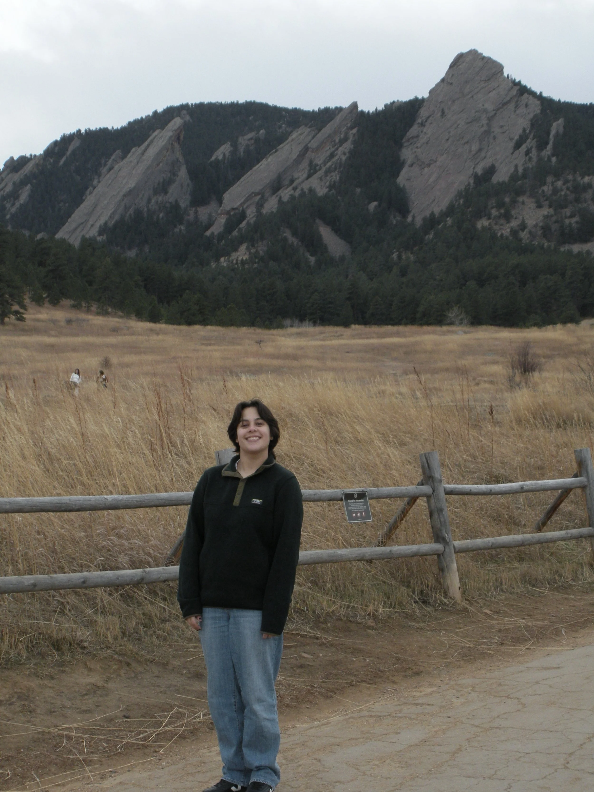 A young woman with short dark hair smiling and standing in a field of tall grass in front of a wooden fence. In the background are mountainous rock formations and a tree-covered hillside.