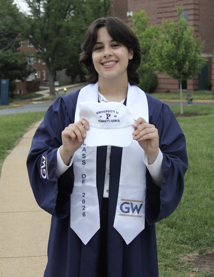 A young woman in a graduation gown holding a white cap with the University of Pennsylvania emblem, standing outdoors on a campus path.