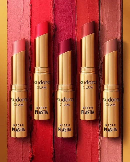 Cinco batons de maquiagem com embalagem dourada, apresentados contra fundos de cores variadas de batom, do vermelho ao nude.