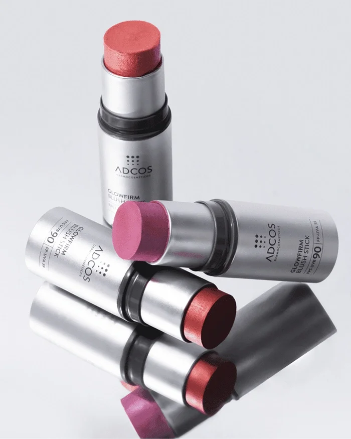 Bastões de blush coloridos em tubos prateados, alguns deitados e outros em pé, sobre fundo branco.