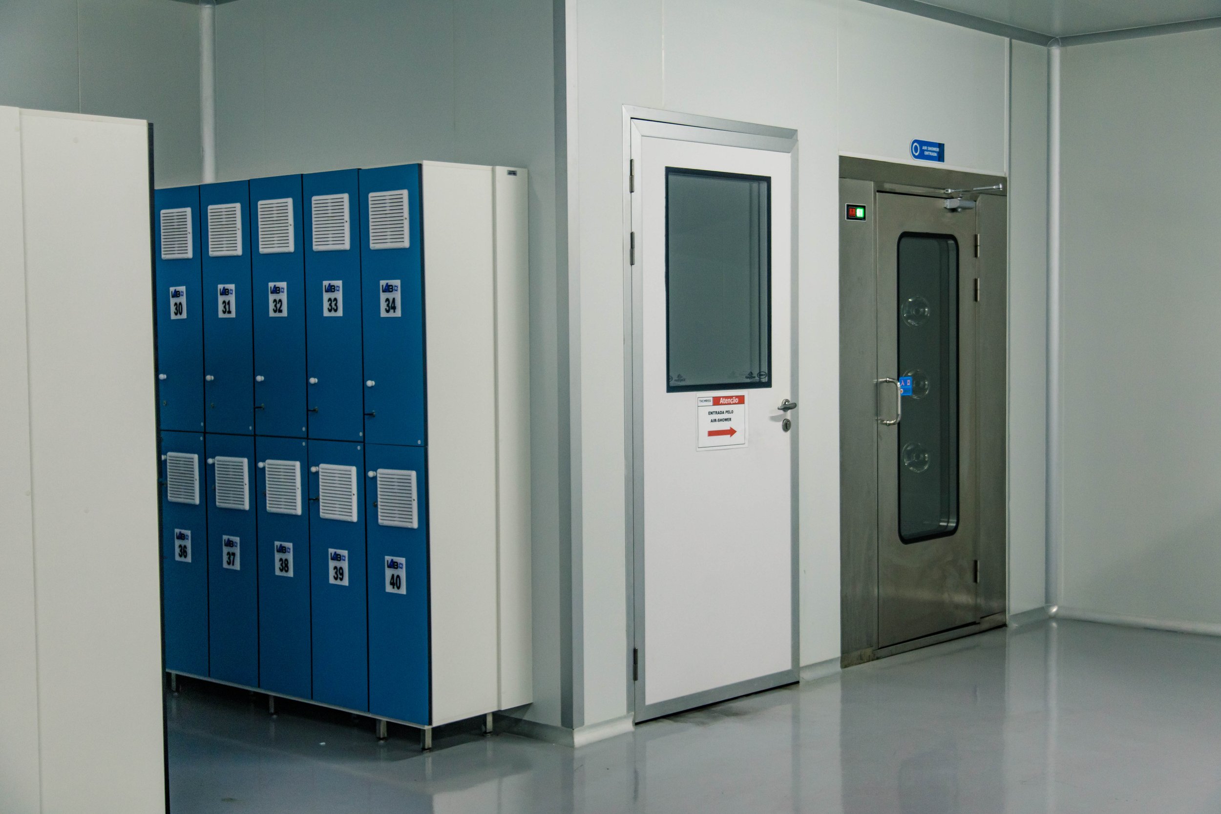 Corredor com armários azuis numerados de 30 a 40 à esquerda e portas de metal à direita, incluindo uma porta de elevador e uma porta de segurança. O ambiente é limpo, com piso de cor clara.