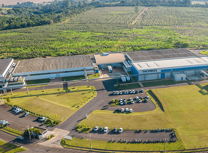 Estrutura industrial com prédios, estacionamento e área verde, ao fundo, plantação agrícola. Fábrica da Thomriss Soluções em Embalagens.