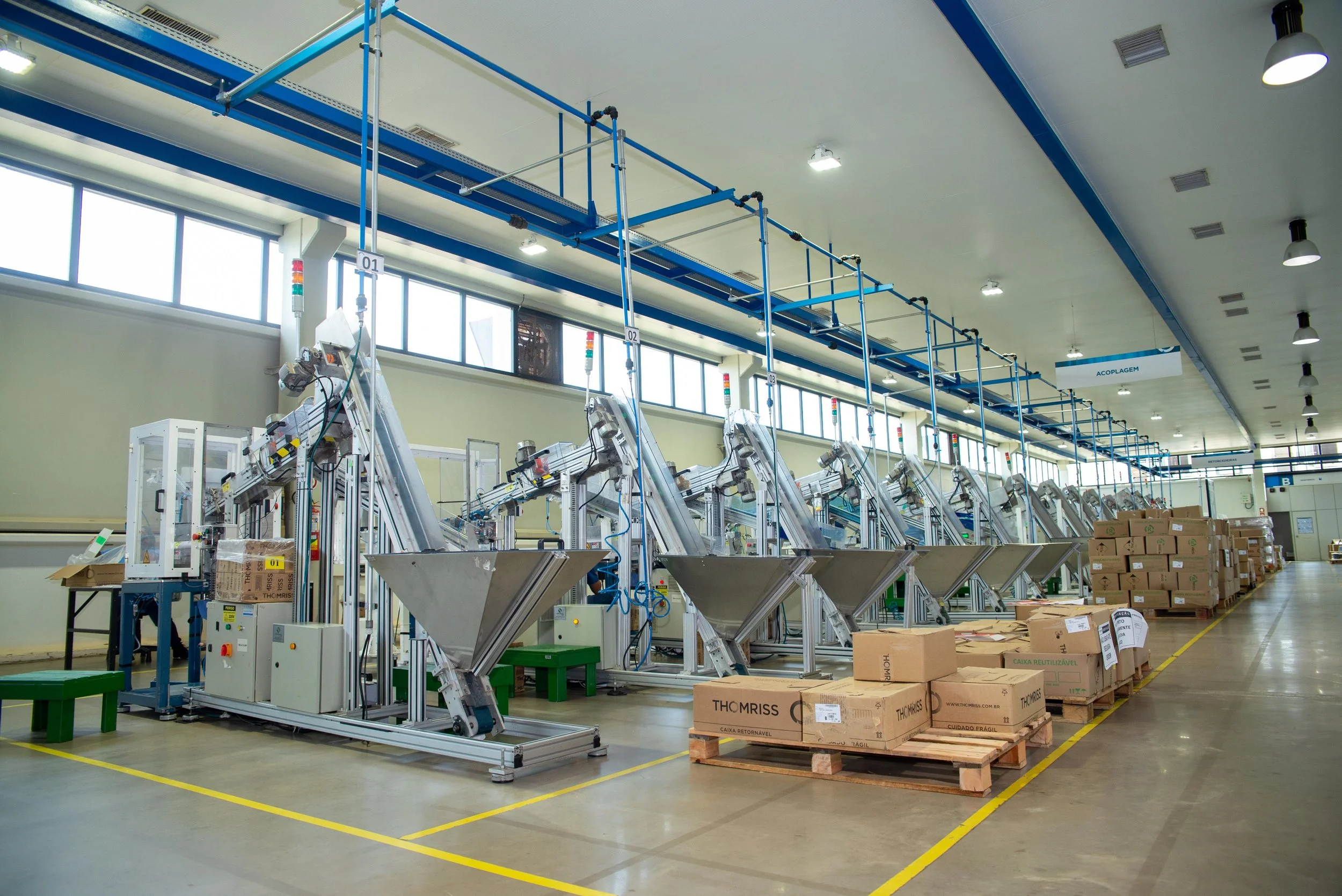 Imagem de uma linha de montagem industrial com máquinas e caixas de papelão, em ambiente de fábrica com iluminação artificial e janelas ao fundo.