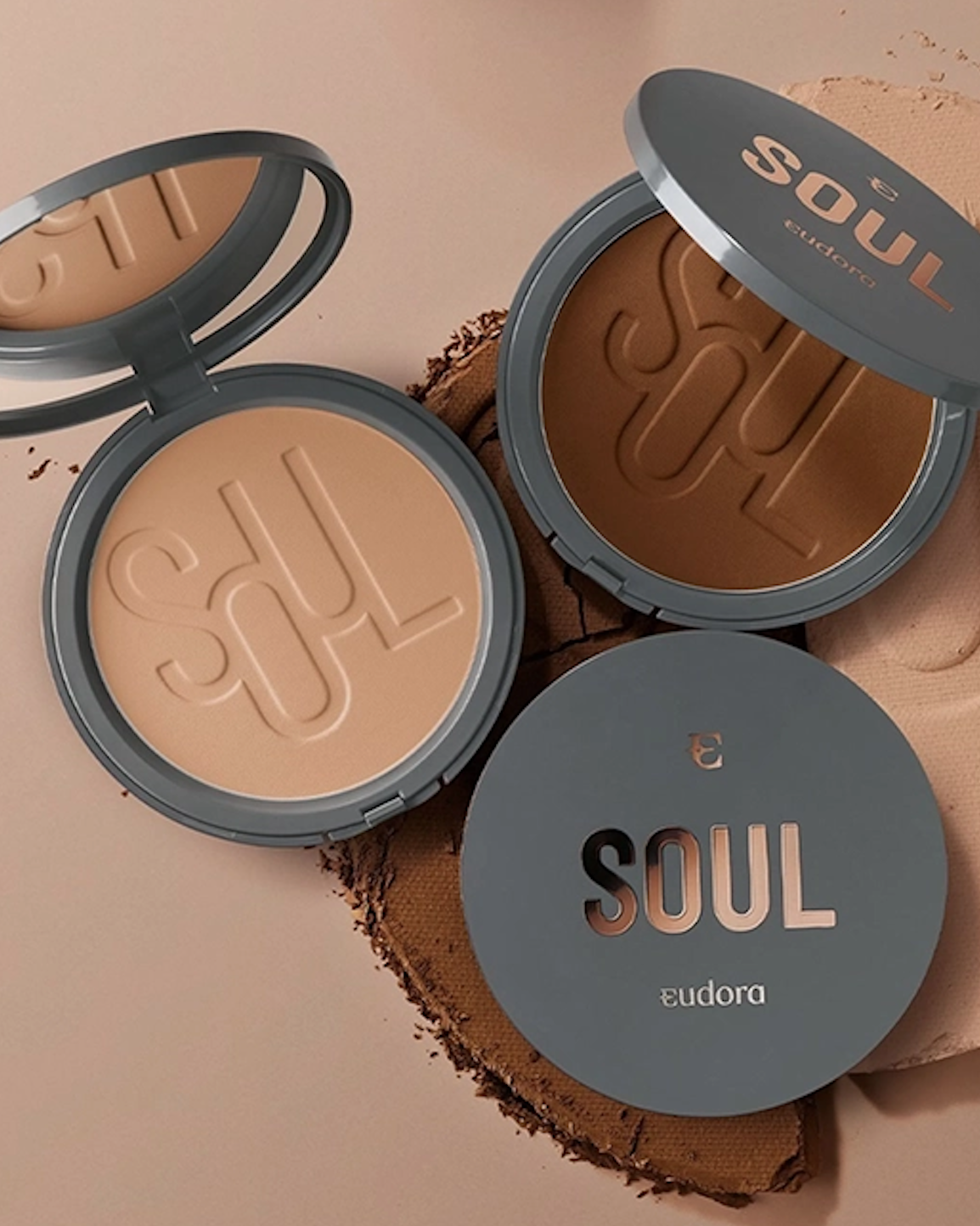 Dois pós compactos da marca Eudora Soul, um aberto revelando a cor bege e o outro fechado com a embalagem cinza e letras douradas, sobre uma superfície de pano bege.