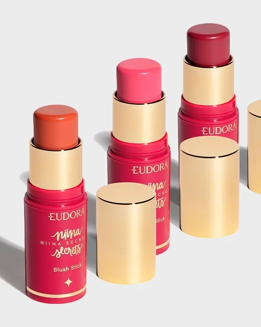 Três paletas de batom em cores diferentes, com embalagens douradas e rosadas, de marcas distintas, exibidas em uma superfície branca com sombras abaixo.