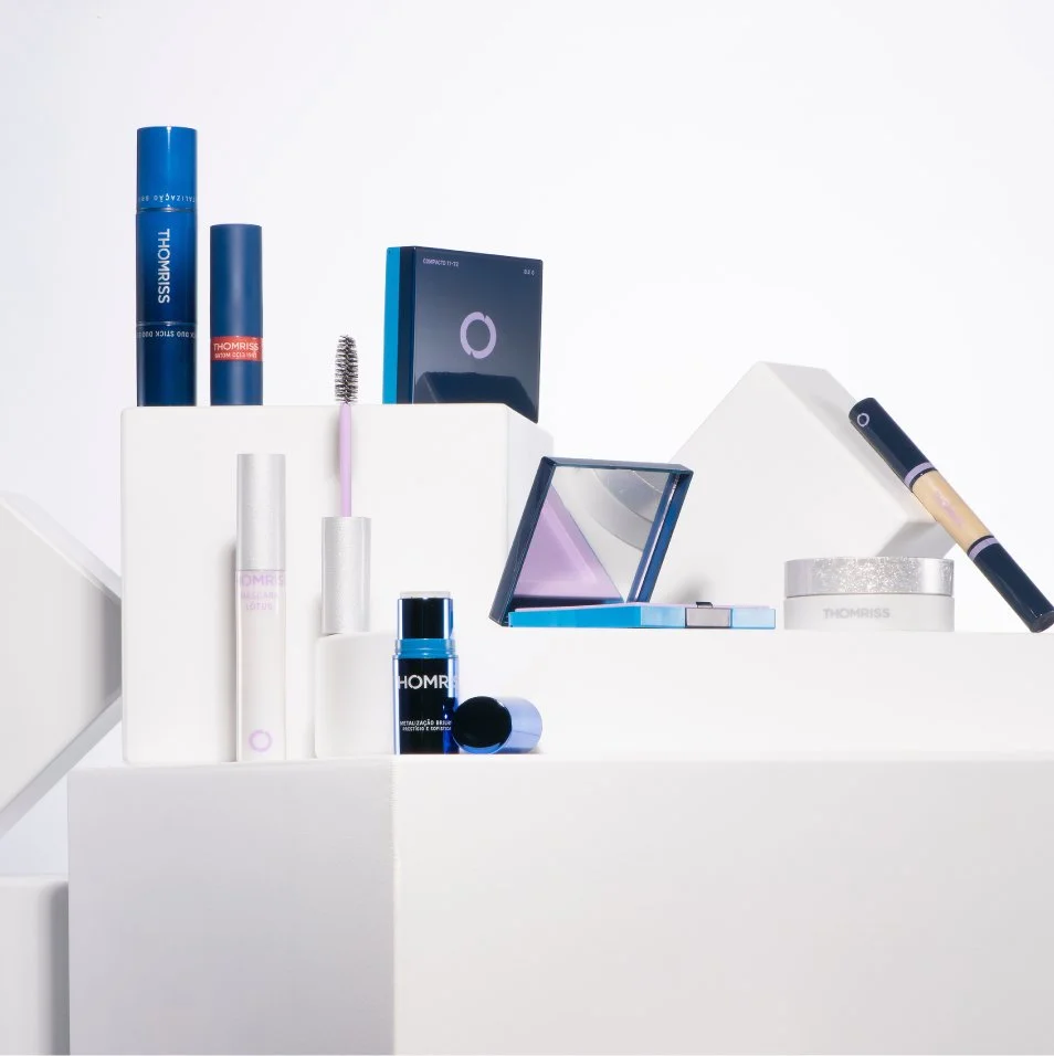 Diversos produtos de maquiagem e cosméticos em uma superfície branca, incluindo batons, sombra, máscara de cílios, base, pó compacto e outros itens de beleza da Thomriss.