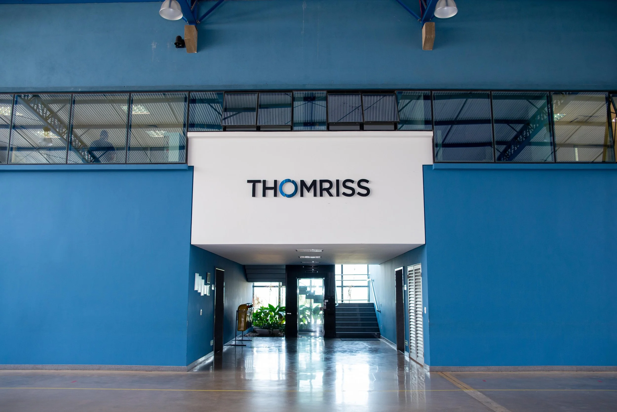 Entrada de um prédio azul com o nome 'THOMRISS' em letras pretas em uma placa branca. Há uma área interna visível com escadas, corredores e plantas, iluminada por luz natural.