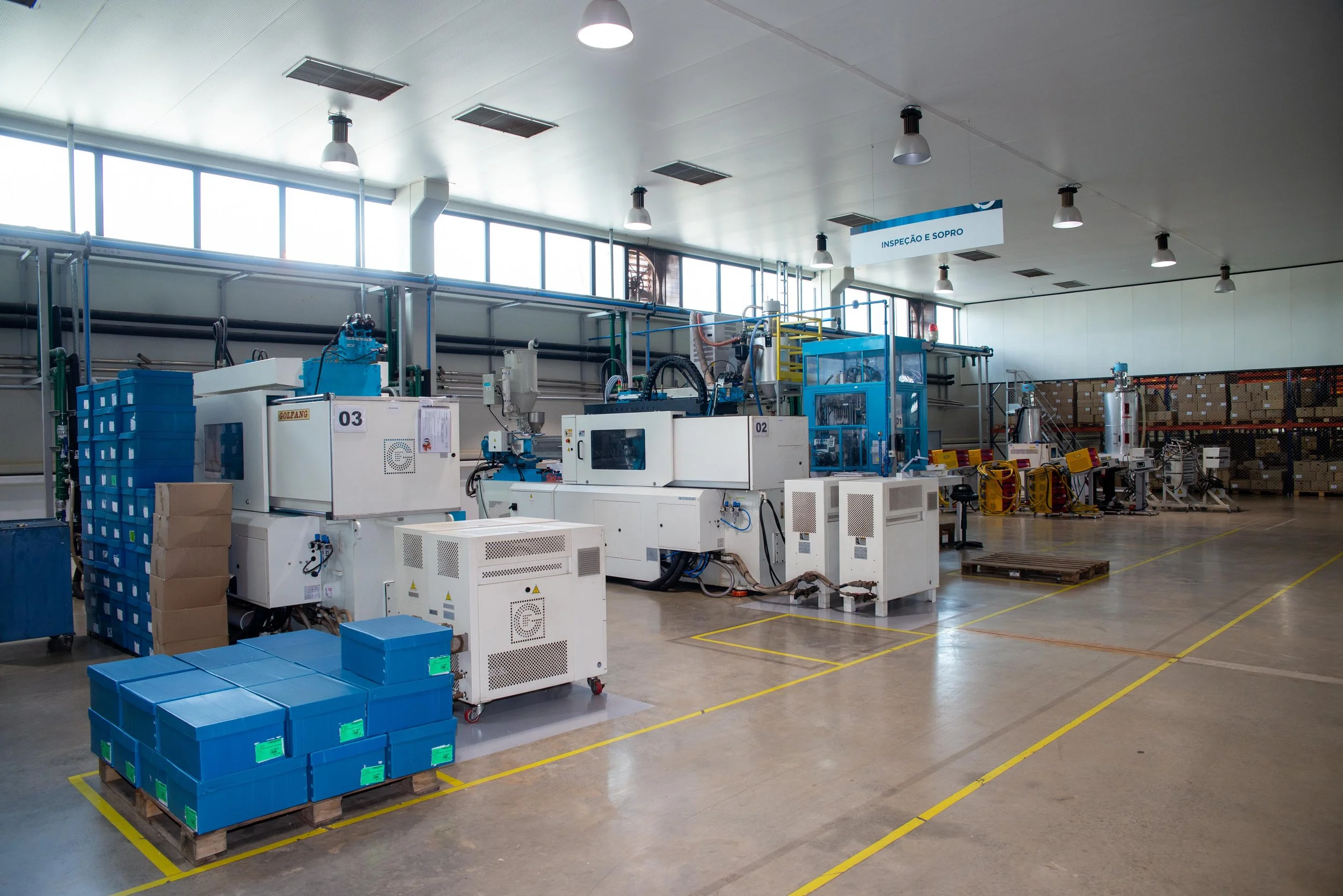 Interior de uma fábrica com máquinas e equipamentos industriais, caixas plásticas azuis e pallets de madeira no chão, com uma placa que diz 'Inspeção e Sopro' suspensa no teto.