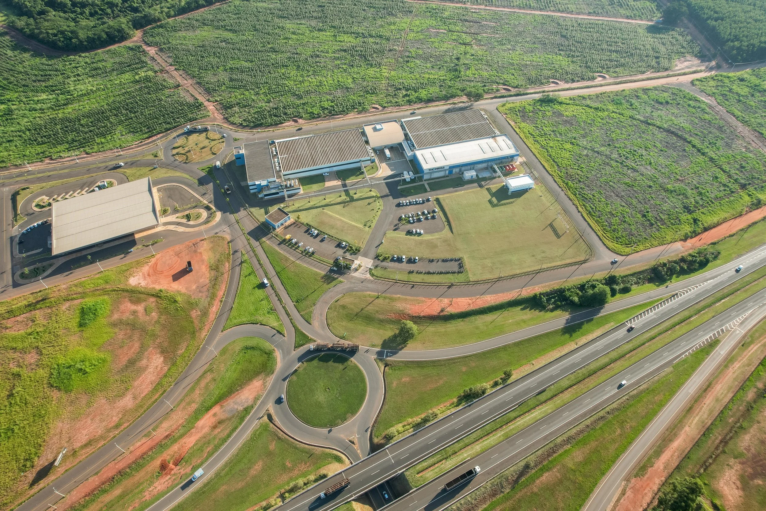 Imagem aérea de uma área industrial rodeada por fazendas e áreas verdes, com ruas, carros e predios vistosos.