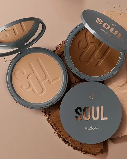 Dois pós compactos da marca Eudora Soul, um aberto revelando a cor bege e o outro fechado com a embalagem cinza e letras douradas, sobre uma superfície de pano bege.