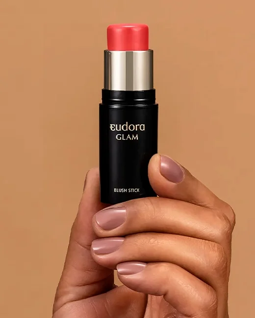 Mão segurando um suporte de blush em bastão da marca Eudora Glam com tom rosa claro.