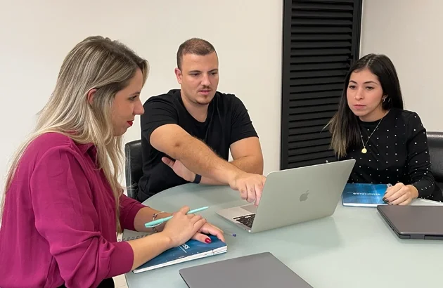 Três pessoas, duas mulheres e um homem, sentados em uma mesa de reunião com laptops e notebooks, em um ambiente de escritório.