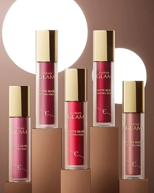 Cinco frascos de batom líquidos Eudora Glam Matte Blur Segunda Pele, variando de tons rosados a avermelhados, sobre suportes em diferentes alturas com fundo de luz e sombra.