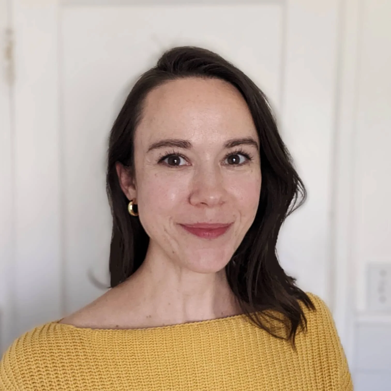 A woman with dark brown hair, wearing a yellow top, and small gold hoop earrings, standing in front of a white door.